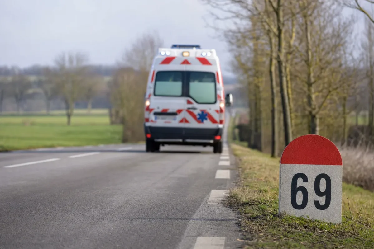 vue de dos d'une ambulance sur une route de campagne rectiligne qui s'etire vers l'horizon, panneau de distance visible